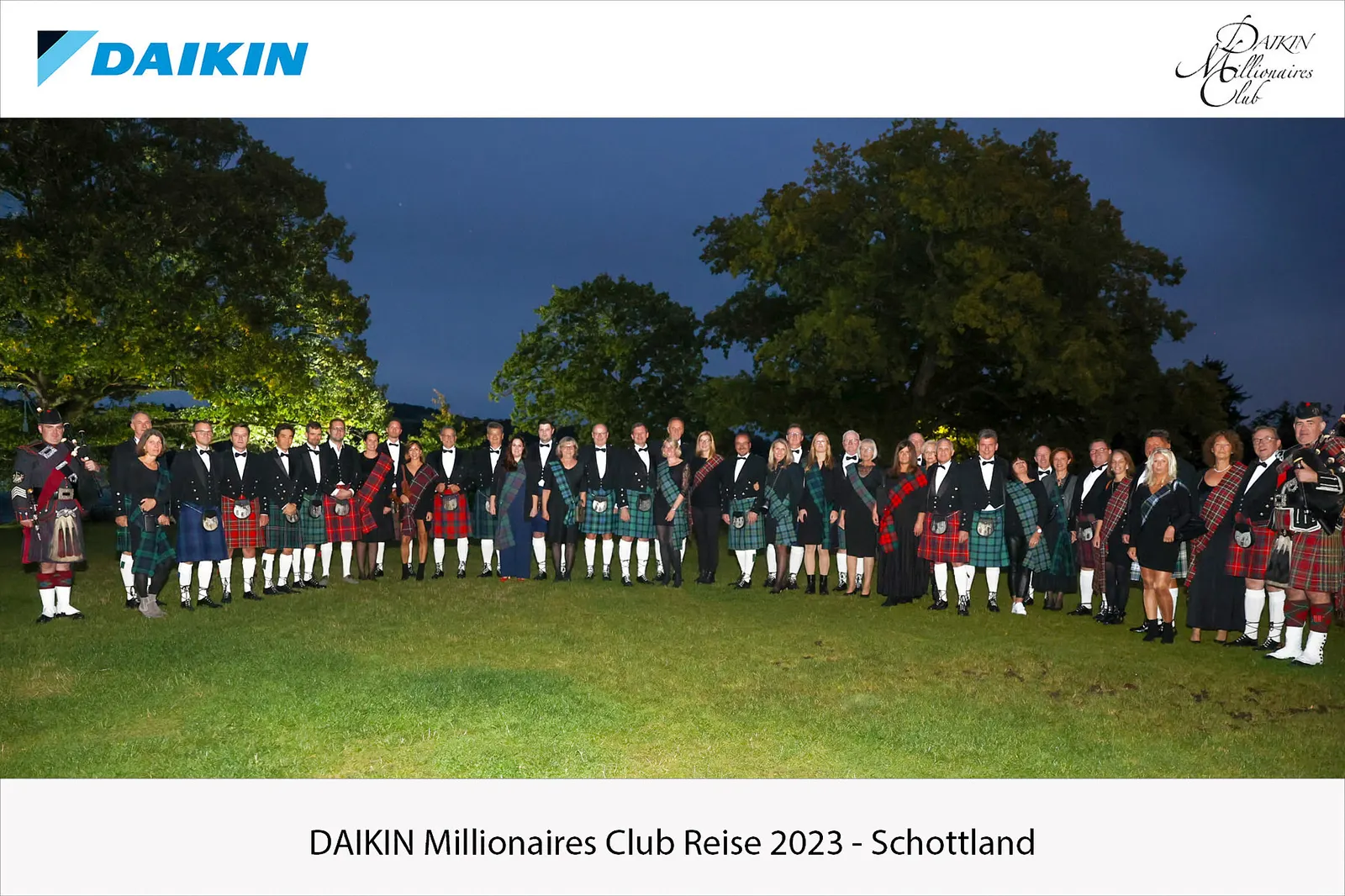 Large group in Highland dress gathered on a lawn at dusk with bagpipers at either end